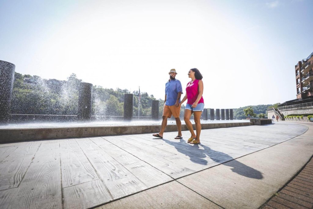 A couple walks hand-in-hand along a waterfront promenade past a fountain