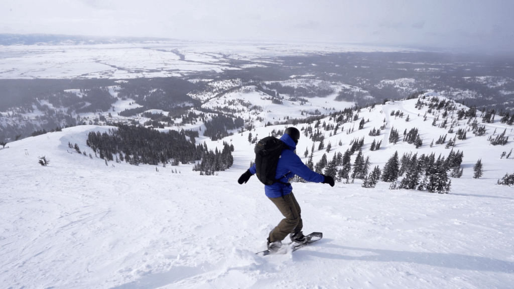 A snowboarder descends a snowy mountain slope with a vast valley in the background