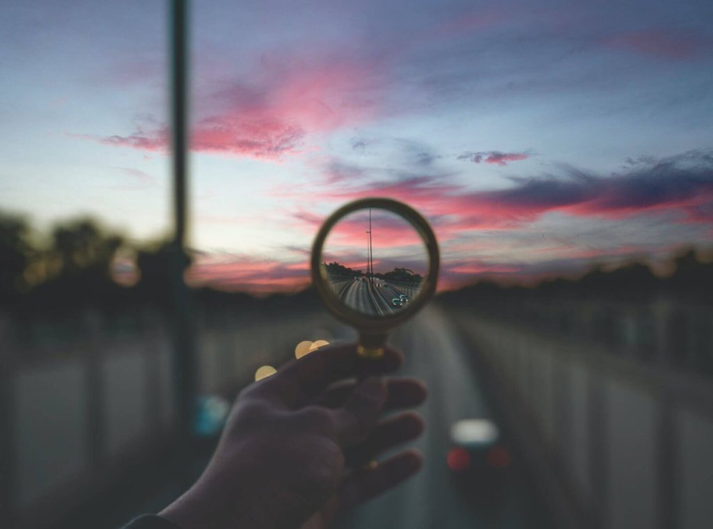 A hand holds a magnifying glass showing a focused view of a highway at sunset