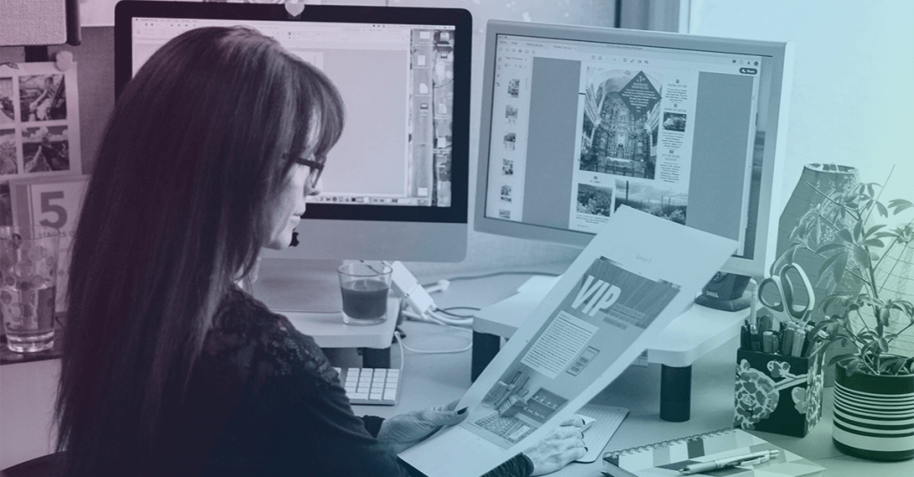A woman working at a desk with two computer monitors and printed documents