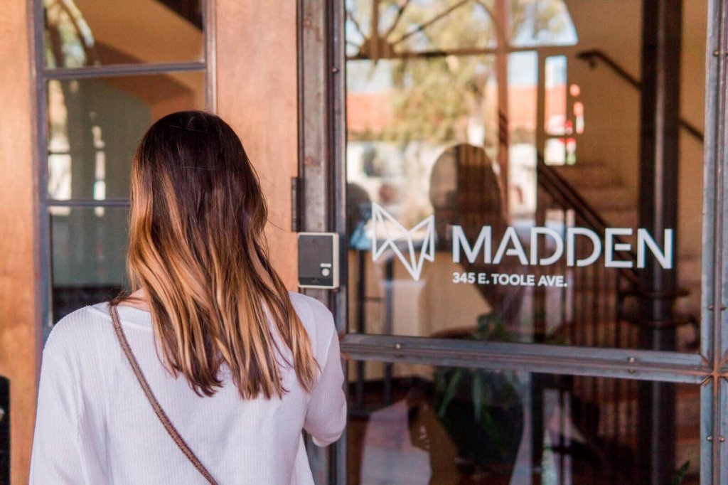 A woman with long hair stands before a glass door labeled Madden