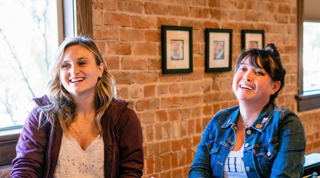 Two women laughing together in front of a rustic brick wall
