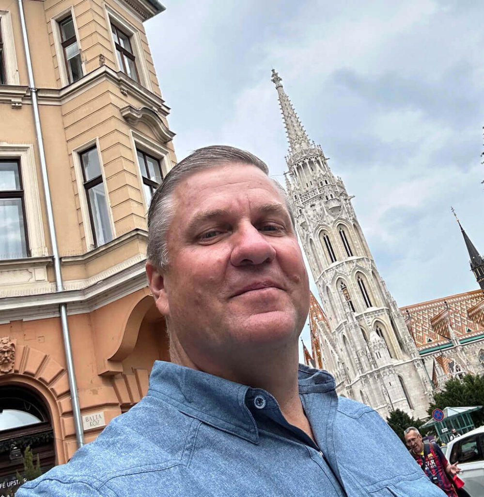 A man in a blue shirt smiles, with a historic building and church in the background