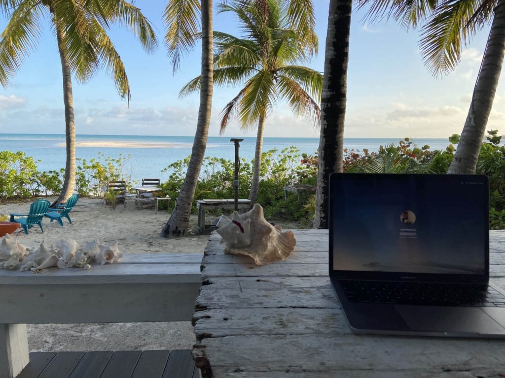 A laptop sits on a rustic wooden table overlooking a tropical beach with palm trees