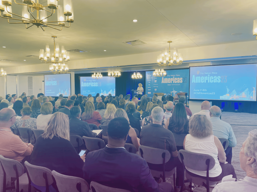 A speaker presents to a large audience at the City Nation Place Americas 23 conference