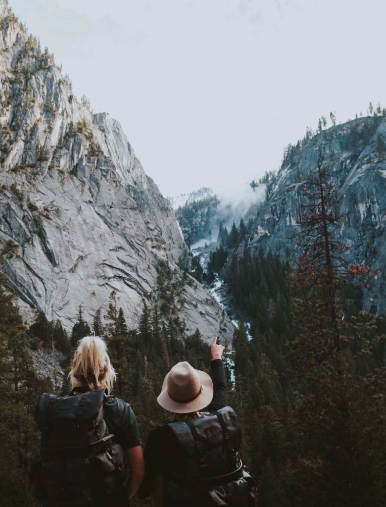 Two hikers with backpacks look at a misty waterfall between large mountain cliffs