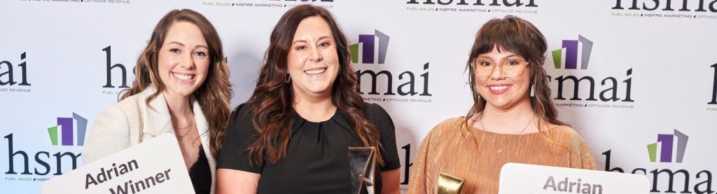 Three women smiling while holding awards in front of a branded backdrop