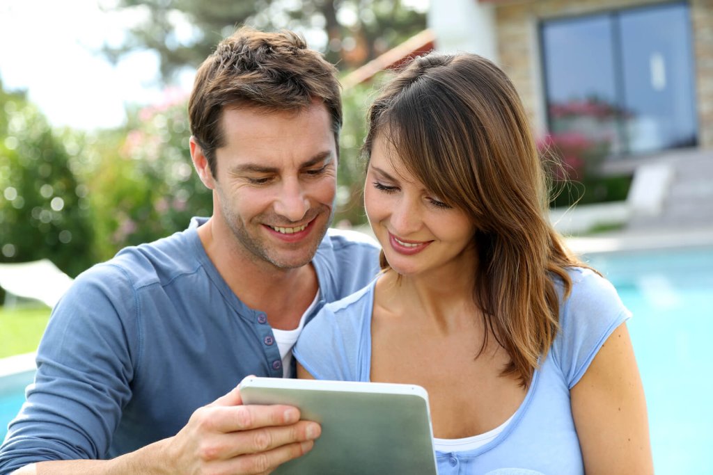 A smiling couple looking at a tablet together outdoors near a swimming pool