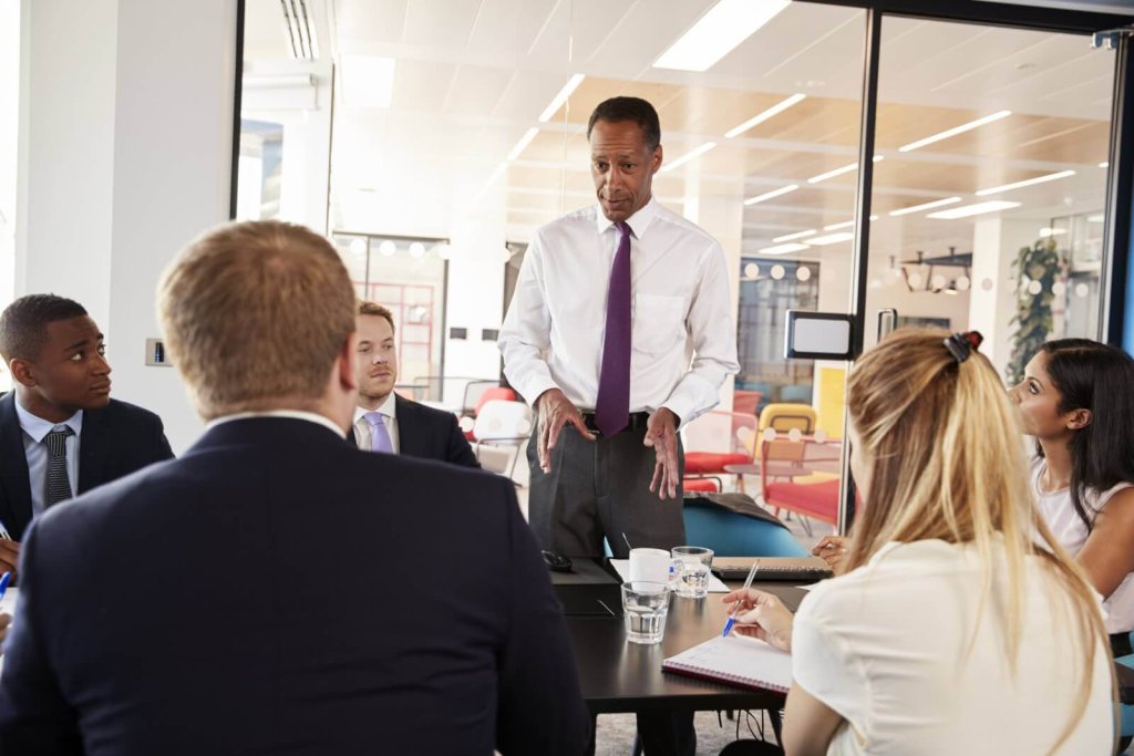 A man in a white shirt and tie leads a business meeting in an office