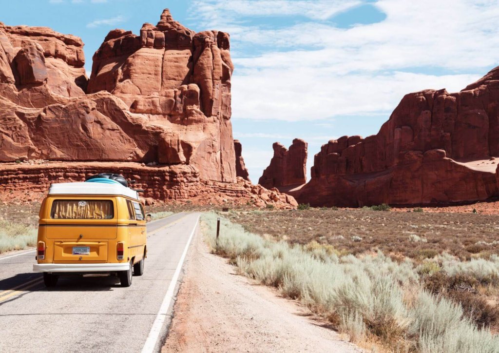 A yellow vintage van driving on a road through a desert landscape with red rocks