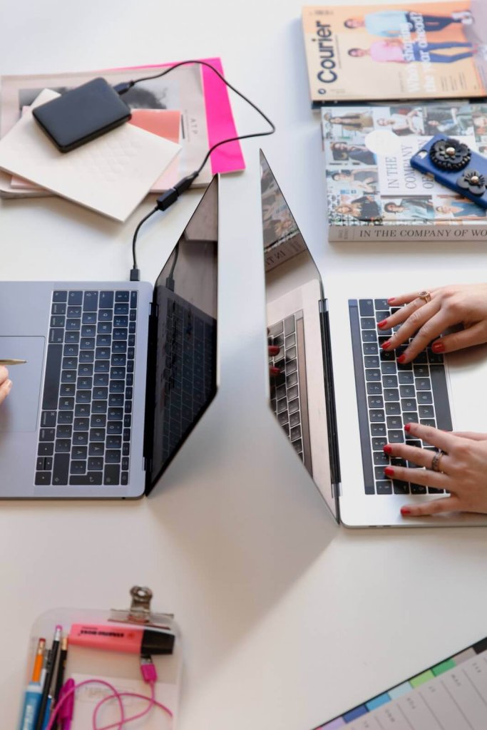 Two people working on laptops on a white desk with office supplies