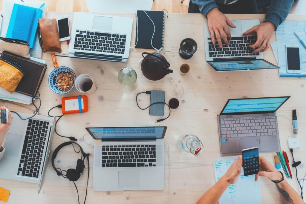 A high-angle view of a wooden table with multiple laptops, phones, and office supplies