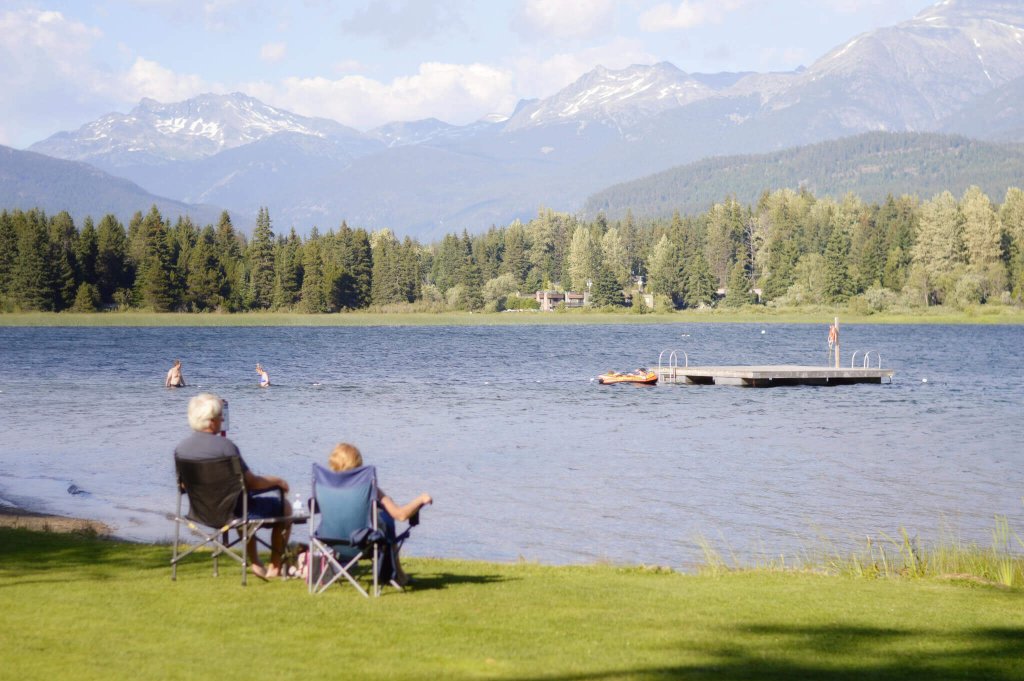 Two people sit in folding chairs on a grassy shore overlooking a mountain lake