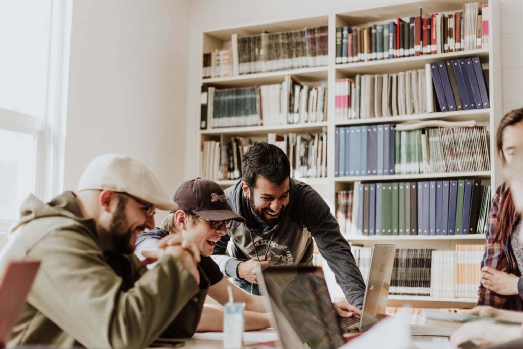 A group of young adults laughing while working together on laptops in a library