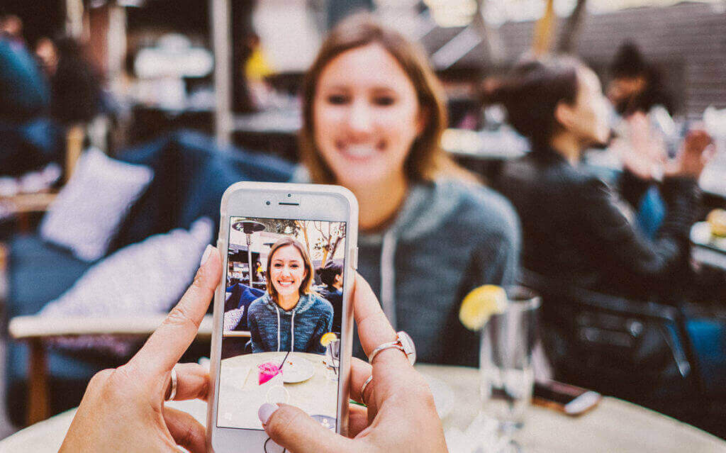 A person taking a photo of a smiling woman sitting at a cafe table