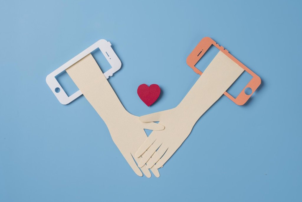 Paper cutout hands reaching out of two smartphones to hold each other with a heart
