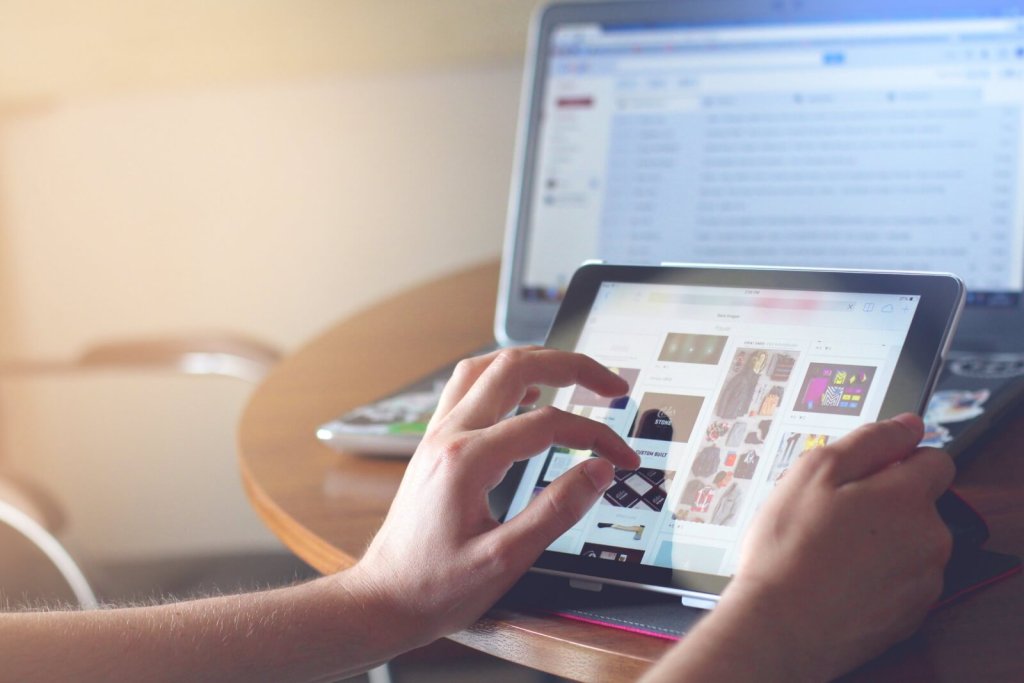 A person using a tablet with a laptop in the background