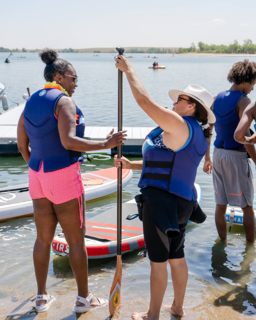 Two women standing in shallow water holding a paddleboard paddle near a dock