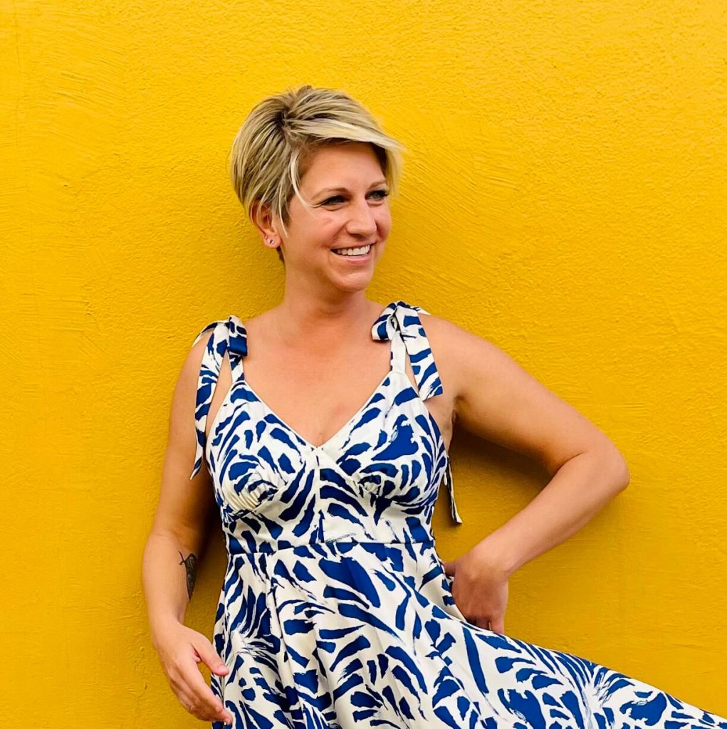 A smiling woman with short blonde hair wearing a blue and white dress against a yellow wall