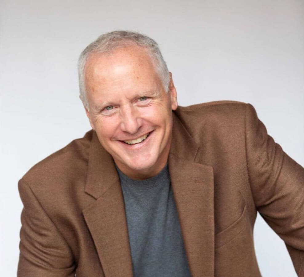 A smiling middle-aged man with grey hair wearing a brown blazer and grey shirt
