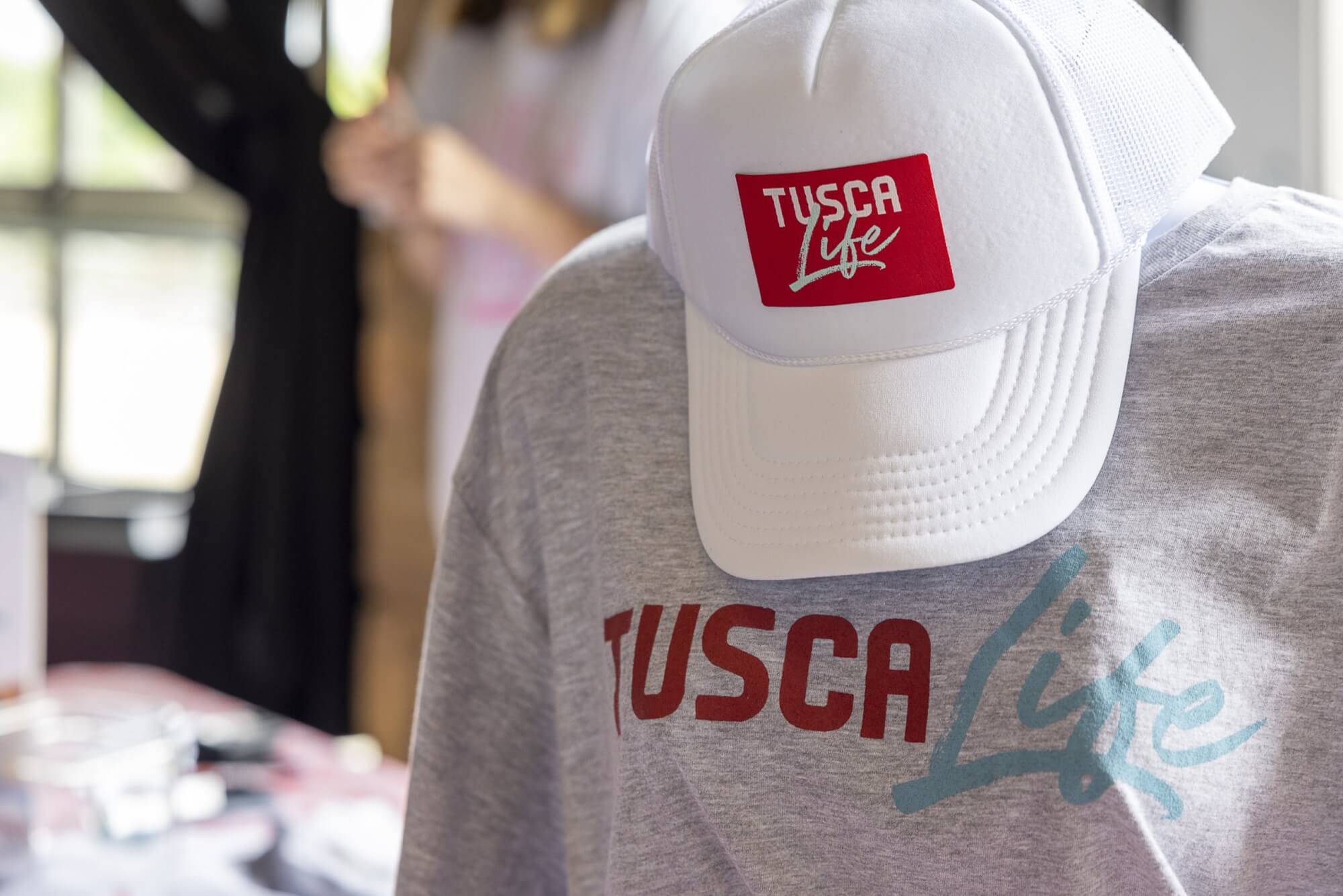 A grey t-shirt and white trucker hat featuring the Tusca Life logo