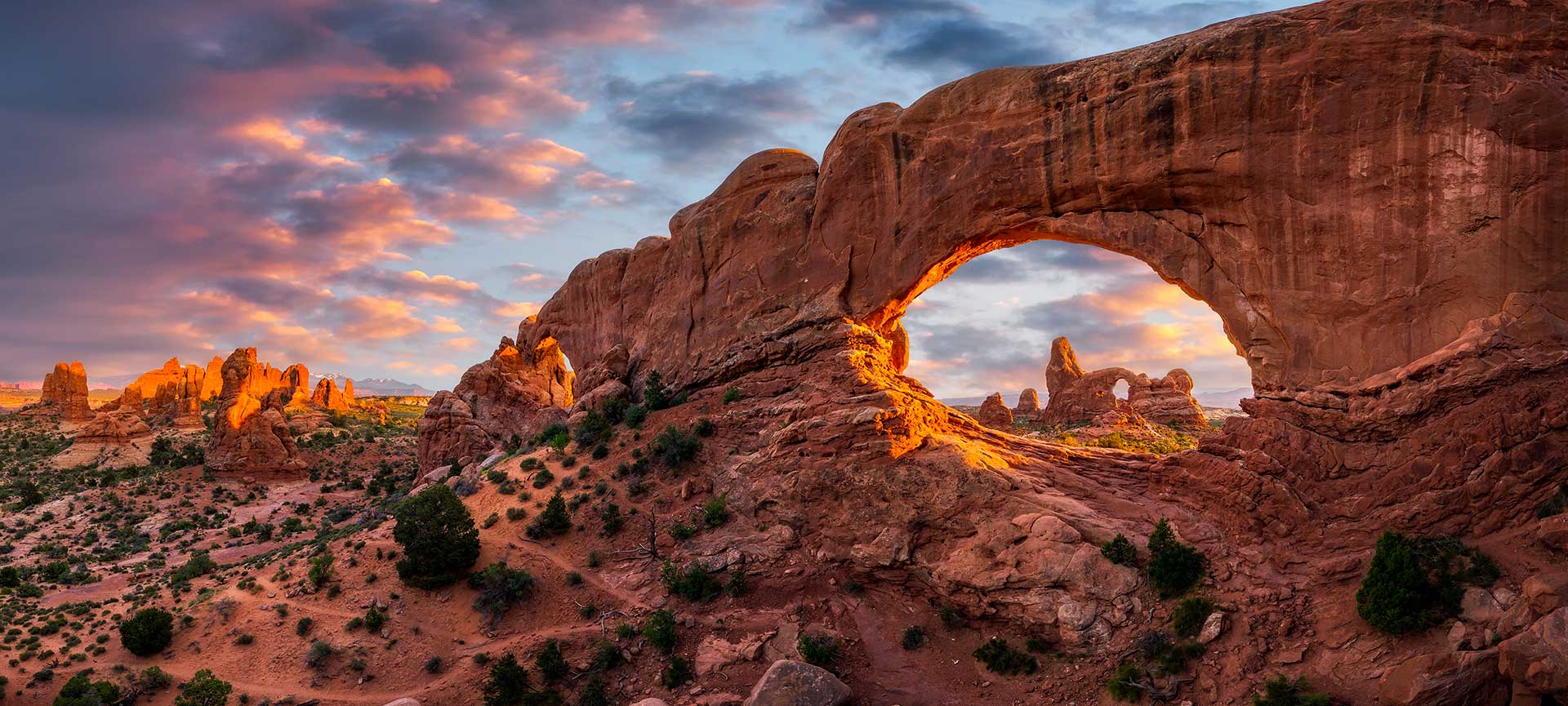 A large natural sandstone arch glows under a dramatic sunset sky