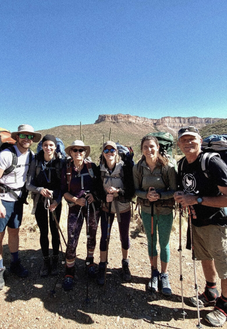 Six hikers with backpacks and trekking poles pose on a sunny trail